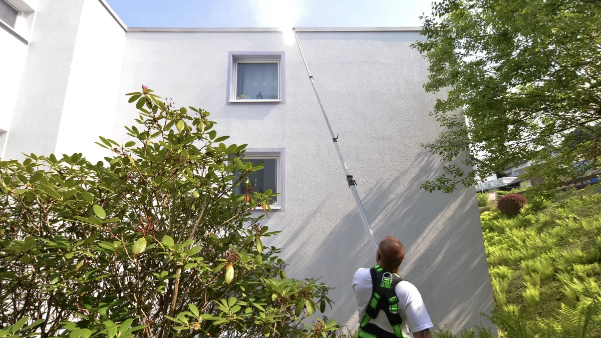 Mann mit Sicherheitsgurt reinigt Hausfassade mit Teleskopstange an weißer Gebäudewand
