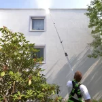 Mann mit Sicherheitsgurt reinigt Hausfassade mit Teleskopstange an weißer Gebäudewand