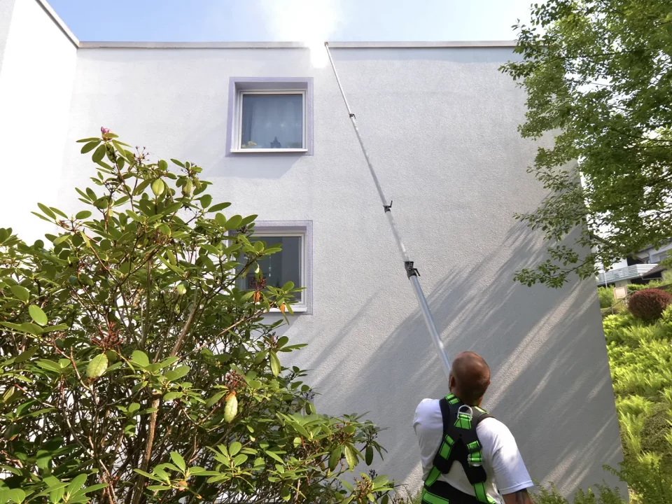 Mann mit Sicherheitsgurt reinigt Hausfassade mit Teleskopstange an weißer Gebäudewand